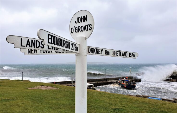 Sign at John O'Groats in Caithness during a storm with waves breaking over the harbour wall